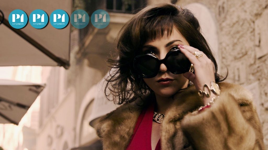 Lady Gaga spelar huvudrollen som Patrizia Reggiani i "House of Gucci". Hon tittar fram bakom ett par svarta solglasögon klädd i brun päls och röd v-ringad topp.