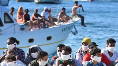 Mercoledì i migranti appena arrivati ​​sedevano in fila sul molo di Lampedusa, mentre passava un battello turistico che trasportava turisti italiani.